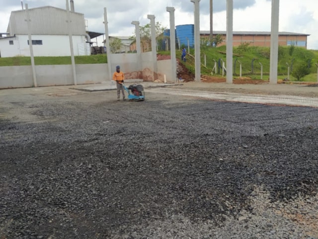Operário utilizando placa vibratória na compactação da base de brita graduada antes da concretagem.